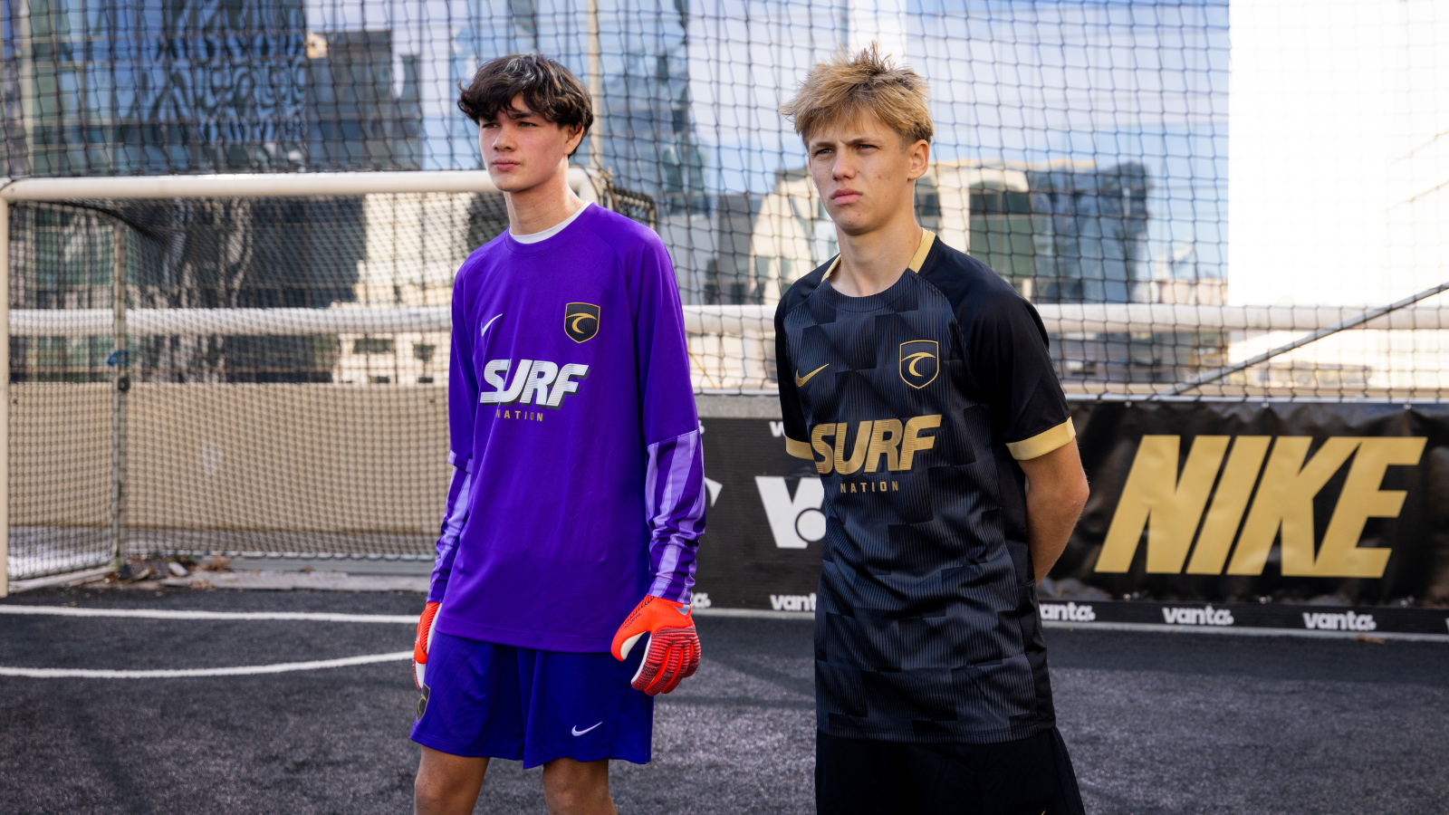 Two teenage soccer players stand on an urban rooftop futsal court in purple and black SURF kits, with a goal and net behind them. Gloves are orange on the player in purple.
