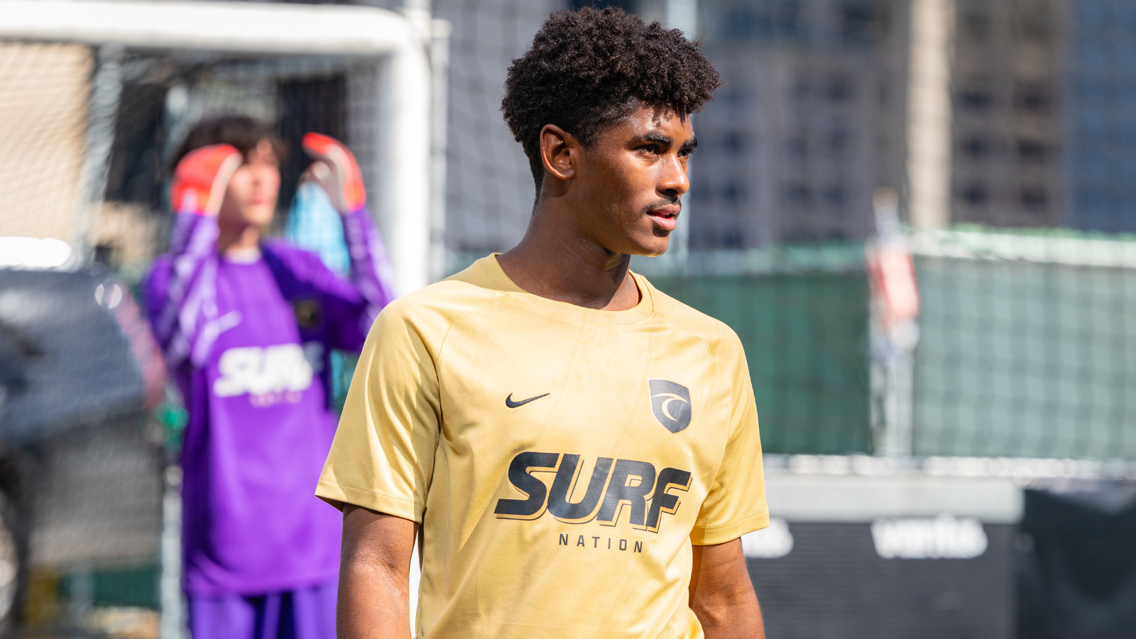Young man in a yellow Surf Nation t-shirt stands on a soccer field, with a goalkeeper in a purple kit blurred in the background behind the goal.