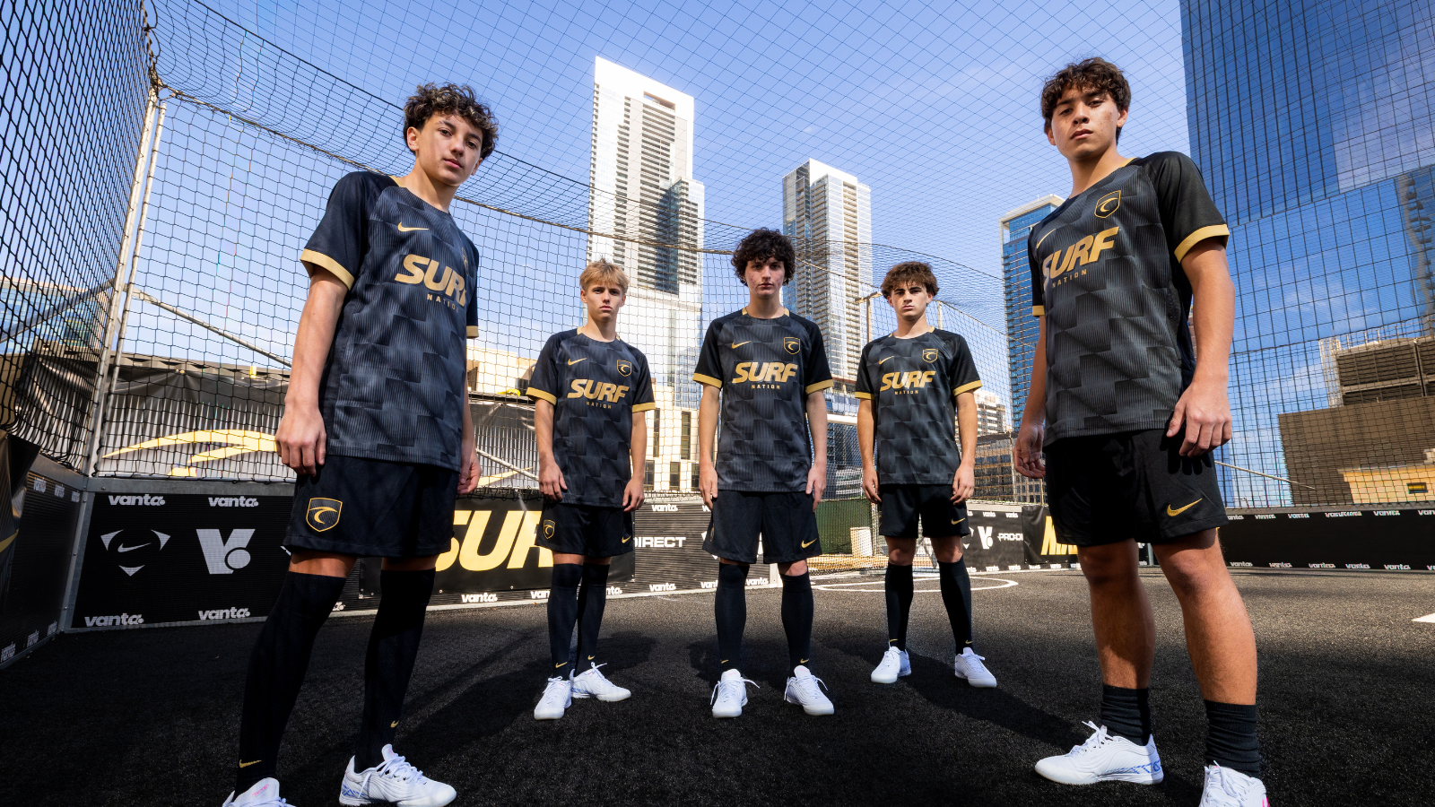 Five young soccer players stand in a line on a fenced rooftop court, wearing black and gold SURF Nation uniforms with city skyscrapers behind them.