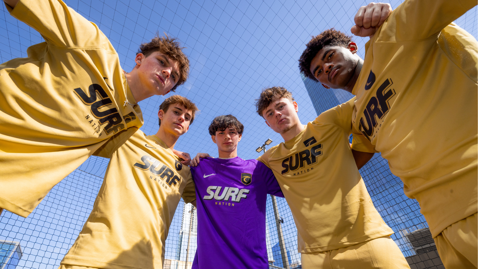 Six young men circle up and lean toward the camera on a fenced rooftop court; five wear gold SURF Nation jerseys, one in a purple goalkeeper jersey, under a clear blue sky.
