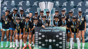 Group of young female athletes in black Champions shirts and caps pose with a tall trophy and a large 'Champions League' bracket board, medals around their necks, in front of an ECNL Playoffs backdrop.