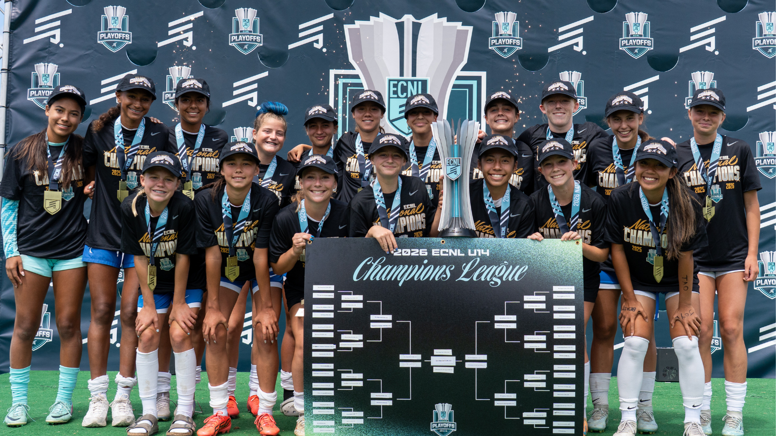Group of young female athletes in black Champions shirts and caps pose with a tall trophy and a large 'Champions League' bracket board, medals around their necks, in front of an ECNL Playoffs backdrop.