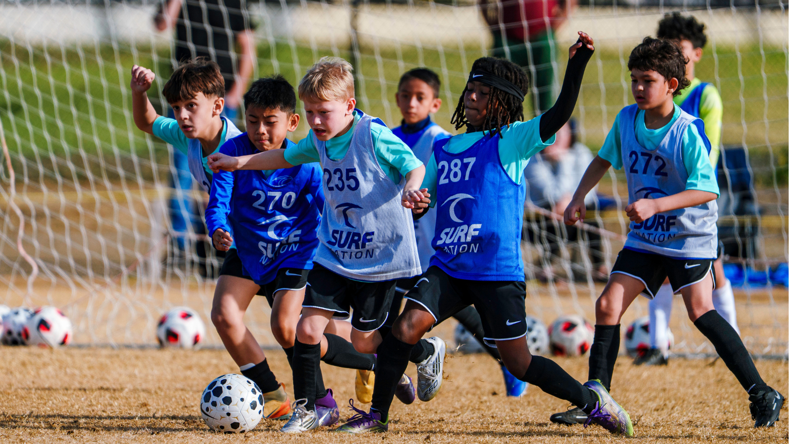 Youth soccer game: several boys chase a ball near the goal on a dry field, wearing numbered bibs (270–287).
