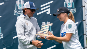 Coach and player exchange a trophy at a sports awards ceremony, backdrop shows ECNL PLAYOFFS logos.