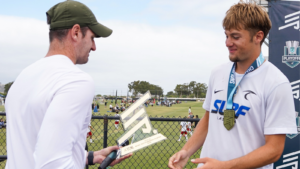 Coach hands a trophy to a smiling young player in a white jersey on a sports field, medal around his neck, by a fence.
