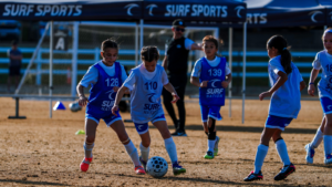 Young girls' soccer match on a dry field as two players battle for a spotted ball while teammates watch in the background under a Surf Sports banner.
