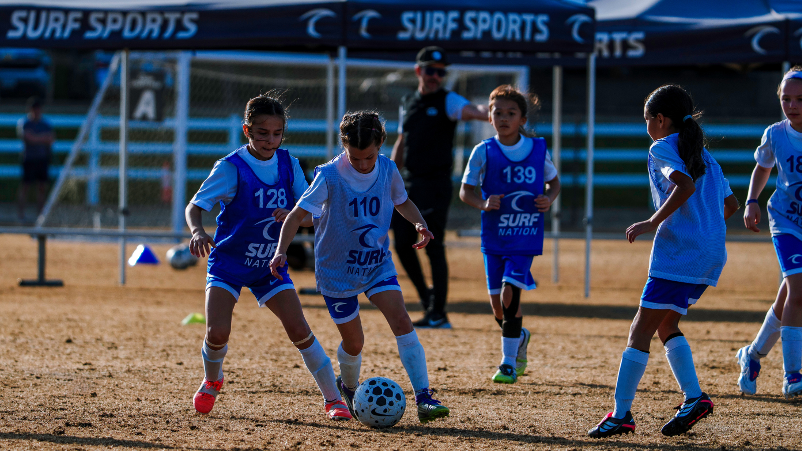 Young girls' soccer match on a dry field as two players battle for a spotted ball while teammates watch in the background under a Surf Sports banner.