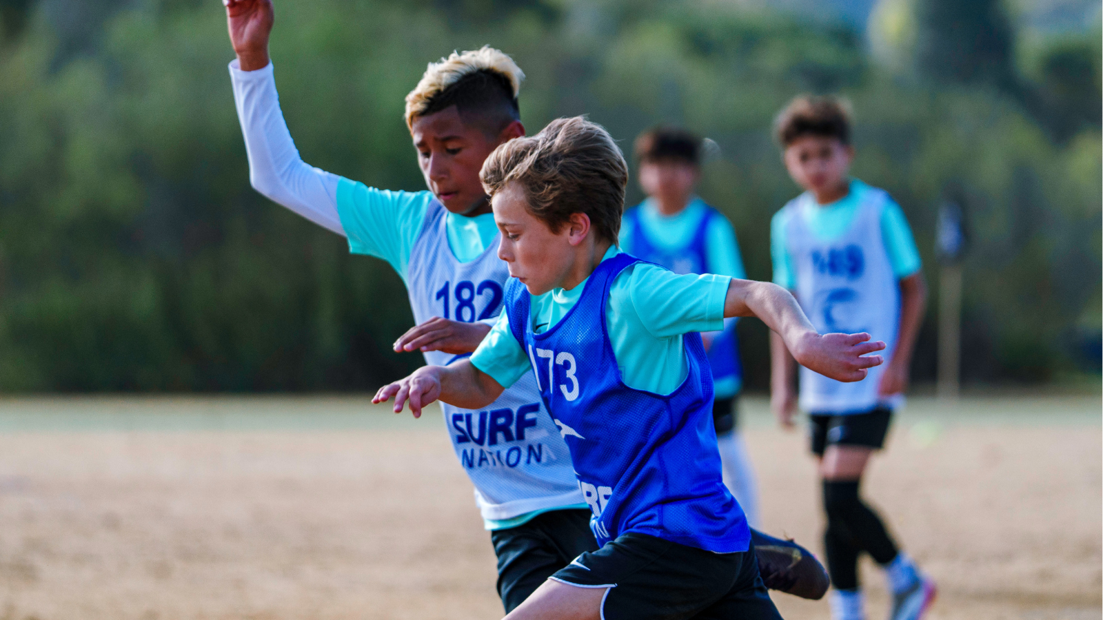 Two young runners in blue bibs sprint on a sandy track, one with arm raised in front. (Informative action)