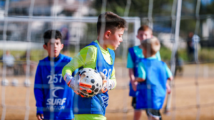 Boy in neon yellow and blue training kit holding a soccer ball inside a goal area during outdoor practice