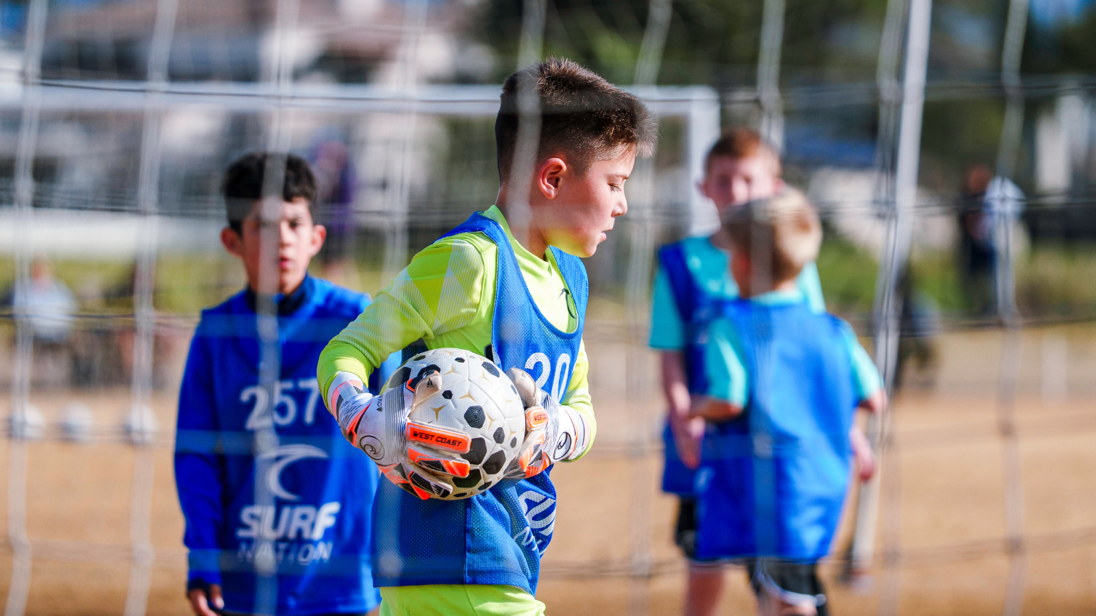 Boy in neon yellow and blue training kit holding a soccer ball inside a goal area during outdoor practice