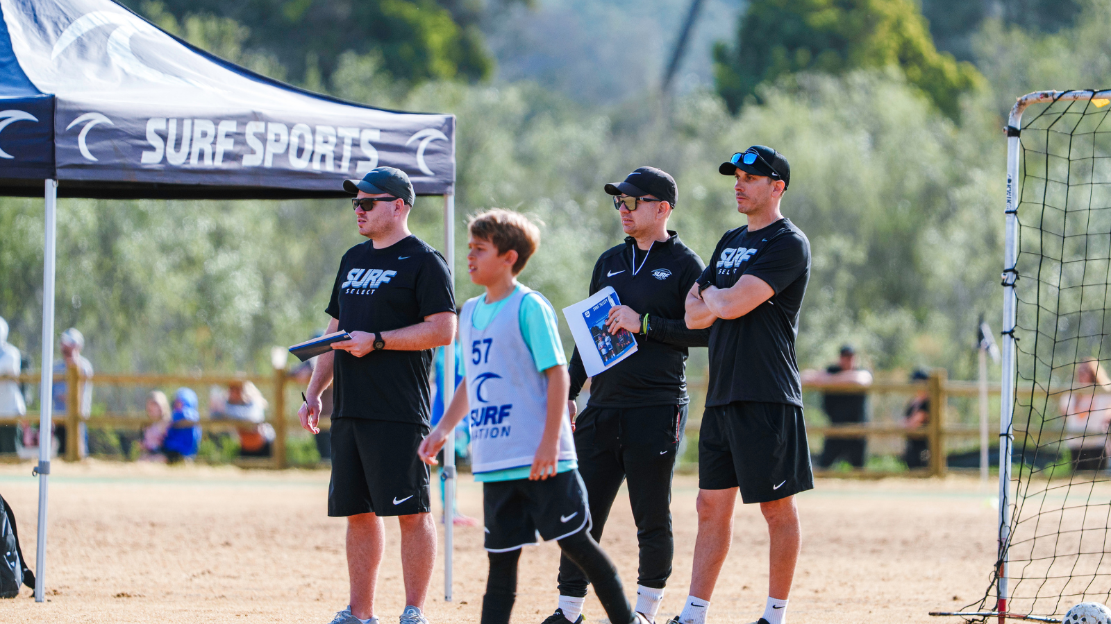 Four coaches and a young surfer with bib 57 stand on a sandy field beside a Surf Sports tent, watching attentively.