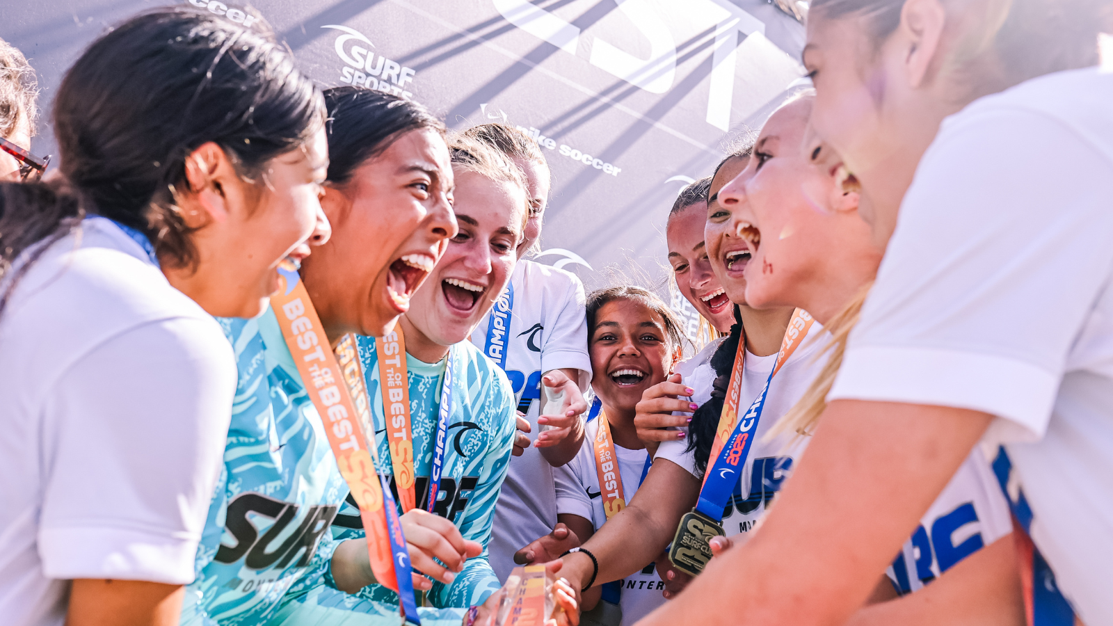 Group of young female athletes in teal jerseys celebrate, smiling and cheering with medals around their necks in a circle.