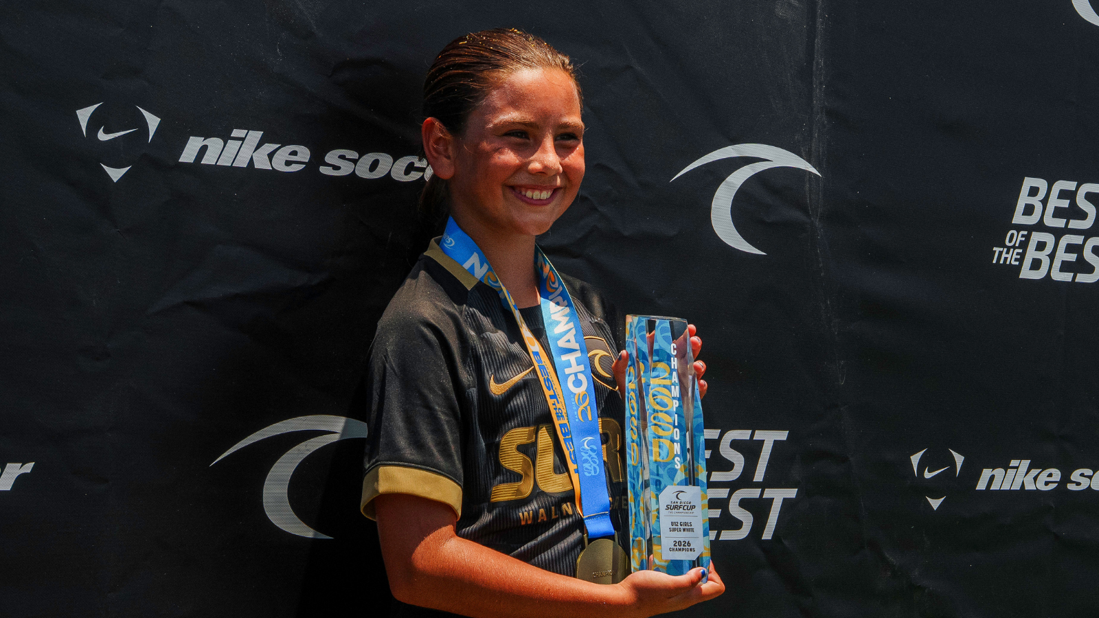 Smiling young girl in a black and gold jersey with a blue medal lanyard, holding a tall blue trophy at a sports event backdrop.
