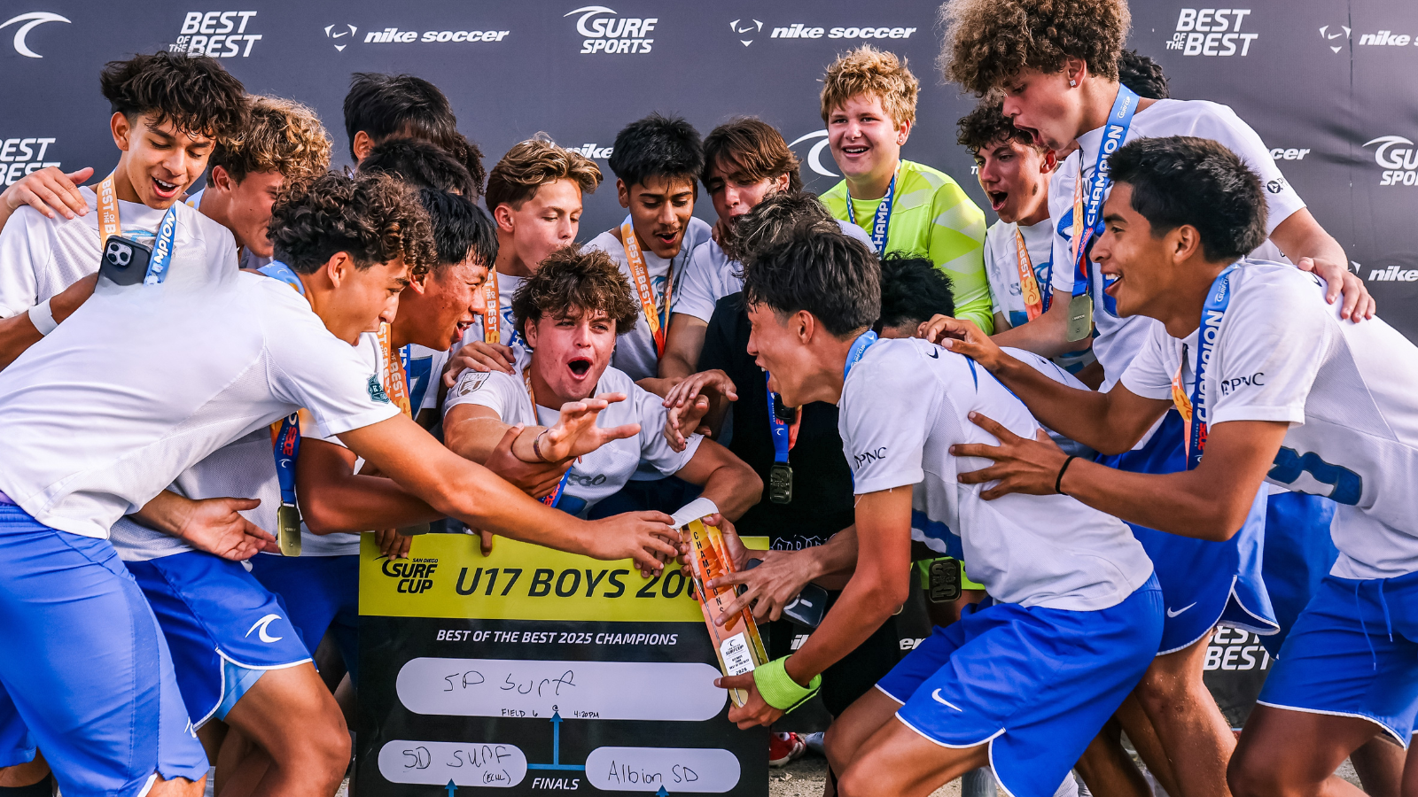 U17 boys soccer team celebrates together holding a trophy after winning the Surf Cup, medals around their necks and a cheering backdrop behind them.