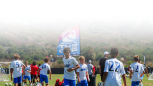 Youth soccer players in white and blue uniforms on a sunny field, with a large banner reading 'WELCOME TO THE CUP' in the background.