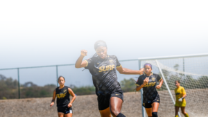 Female soccer player in a black jersey sprinting toward the camera on an outdoor field, with teammates and a goal behind her.