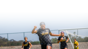 Female soccer player in a black 'SURF' jersey runs toward the camera with a raised fist on an outdoor field; teammates and a goal are in the background.
