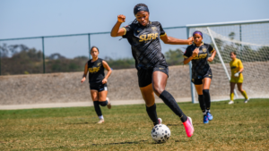 Female soccer player in a black SURF kit sprints toward the ball on a sunny field, teammates in the background and a goal nearby