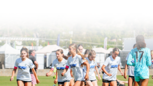 Group of female soccer players jogging on a sunny field in white uniforms; goalie in turquoise on the right.