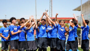 Youth soccer team in blue uniforms celebrate victory, lifting a trophy together on the field. In the background is a goal net and sunny sky.